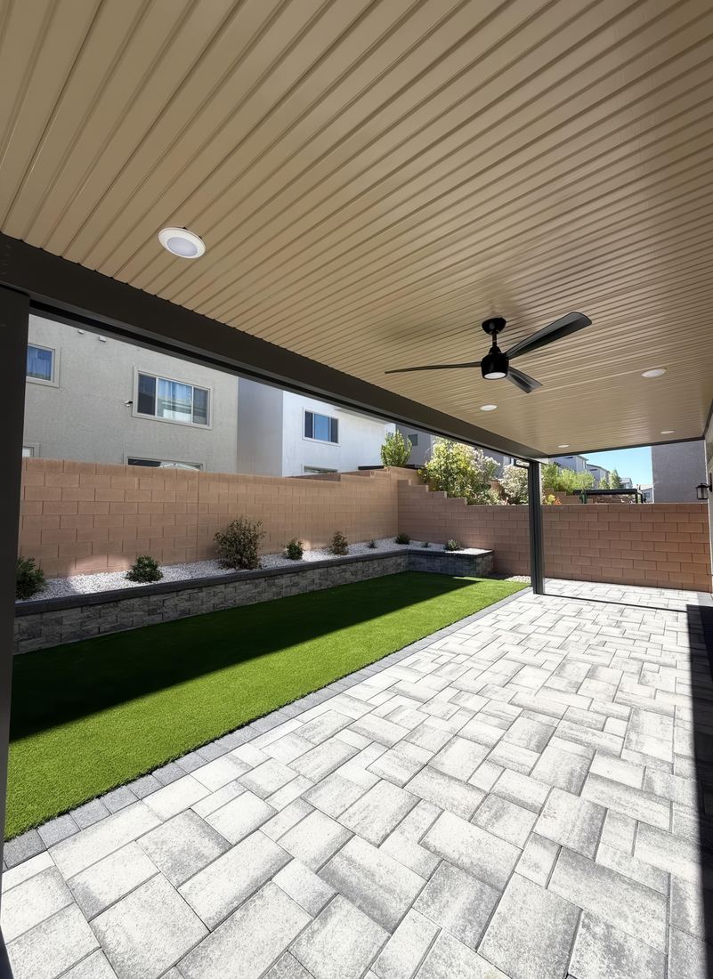 Covered patio with a ceiling fan opening onto herringbone pavers, artificial turf, and a raised rock planter